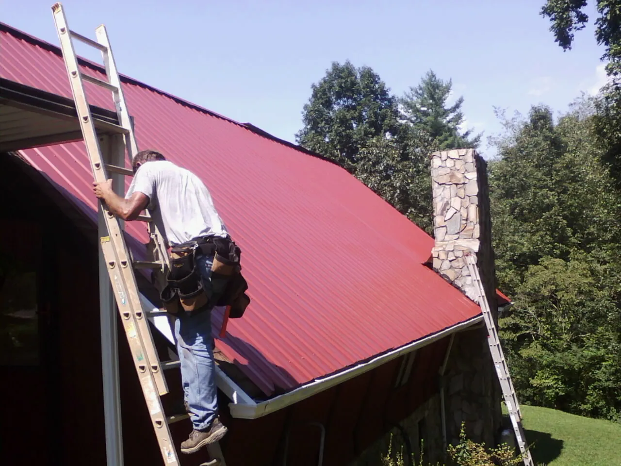 Roofer on a ladder beside a red metal roof and stone chimney — Soffit & Fascia Repair by Roofing Brothers in Bicknell